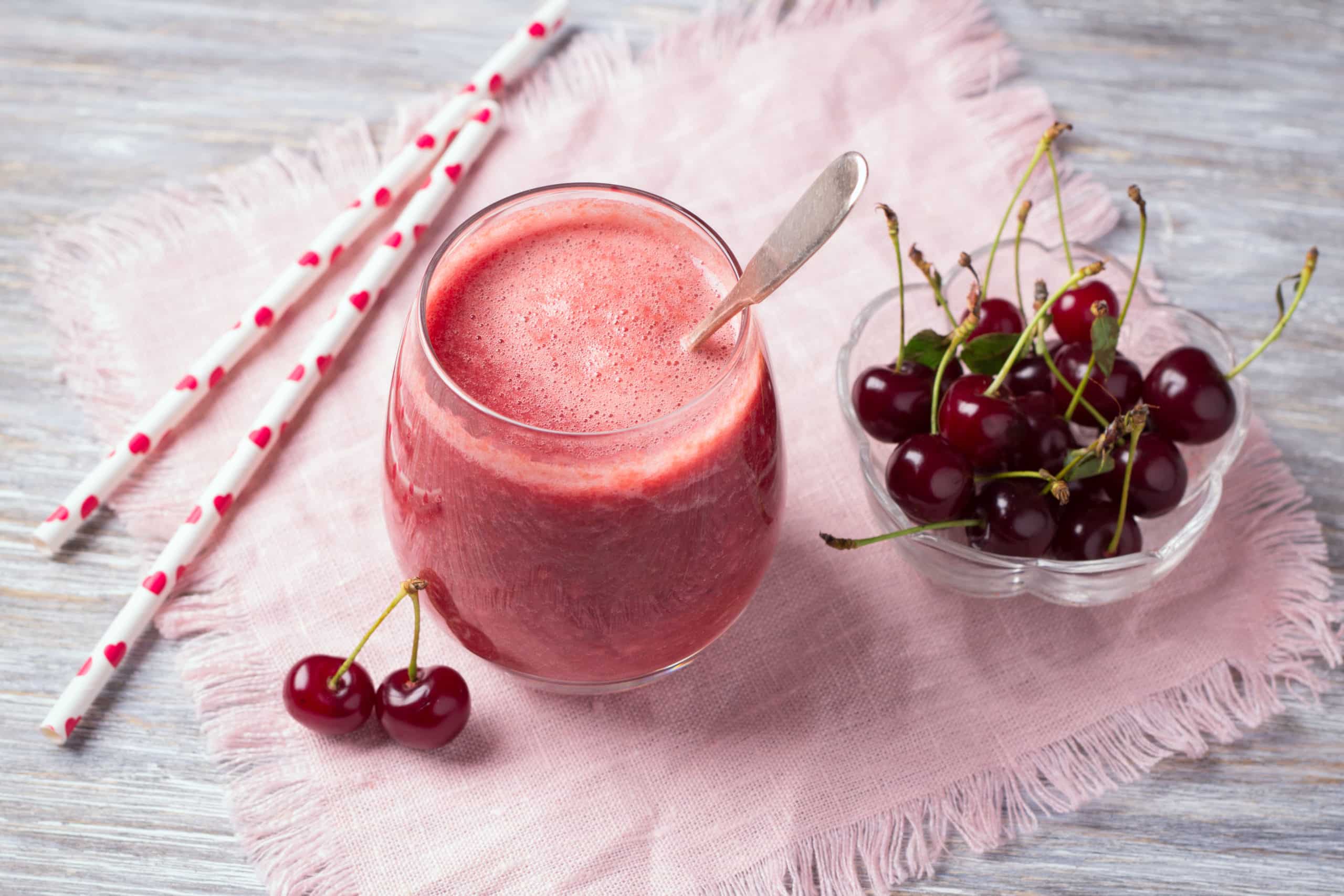 Cherry smoothie in a glass jar with straws and a bowl of cherries next to it