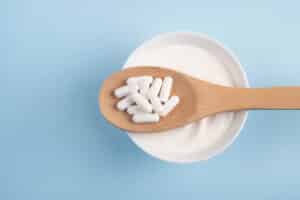 Capsules probiotic on a wooden spoon and bowl with homemade yogurt on light blue background.