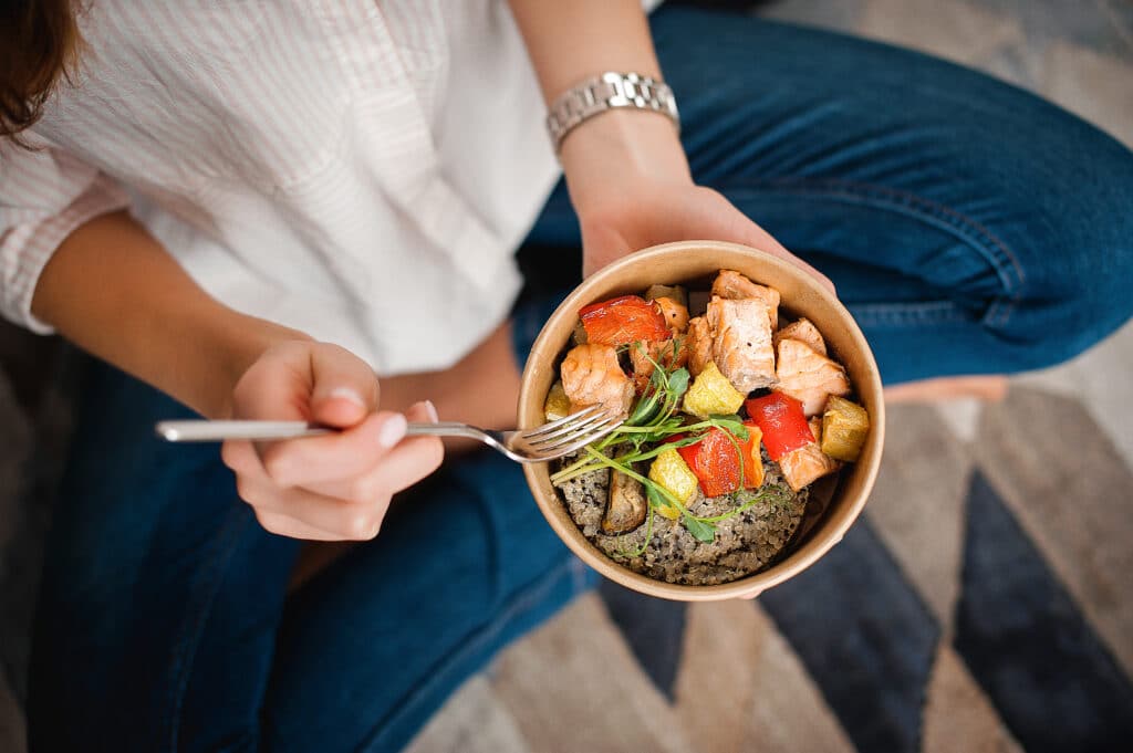 woman sitting in blue jeans enjoying bowl of salmon, rice and colorful veggies