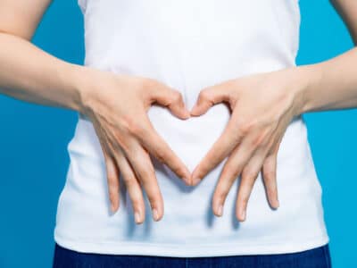 young woman in white shirt who makes a heart shape by hands on her stomach on blue background