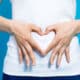 young woman in white shirt who makes a heart shape by hands on her stomach on blue background