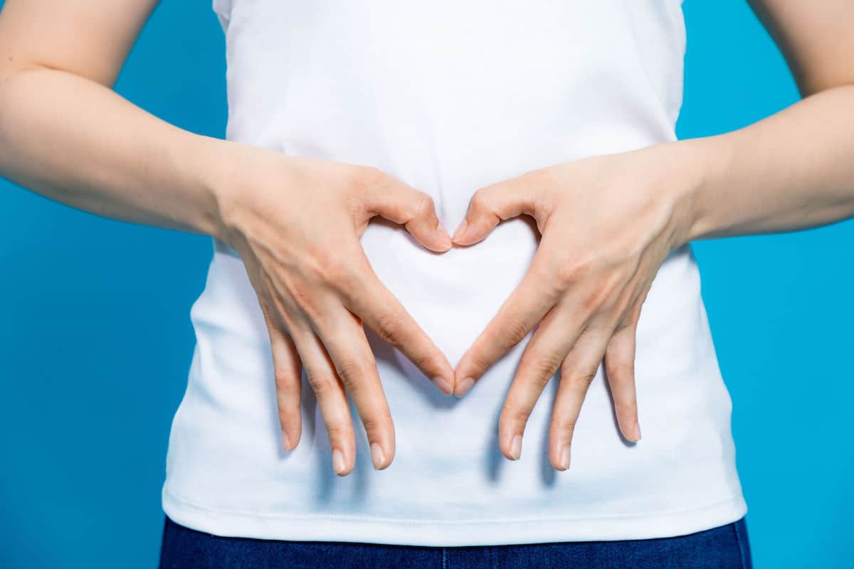 young woman in white shirt who makes a heart shape by hands on her stomach on blue background